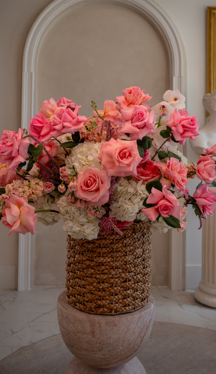 Bouquet of pink and white flowers in a woven basket against a classical interior background.