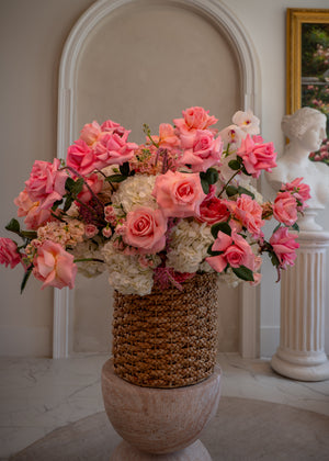 Bouquet of pink and white flowers in a woven basket against a classical interior background.